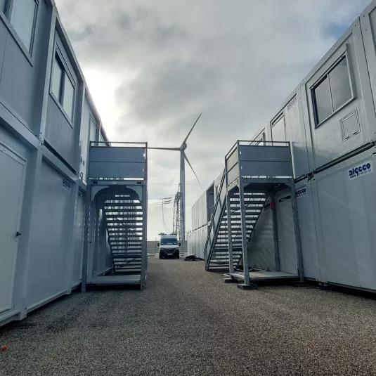 Allée entre bâtiments modulaires ALGECO® à deux niveaux avec escaliers extérieurs et éolienne en arrière-plan
