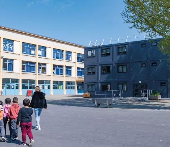 groupe d'enfants et un adulte marchant ensemble sur une grande cour extérieure vers deux bâtiments scolaires