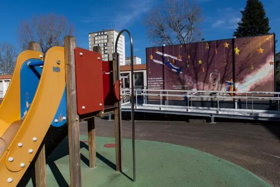 Cour de récréation avec toboggan devant une école modulaire Algeco illustrée d'une citation du Petit Prince
